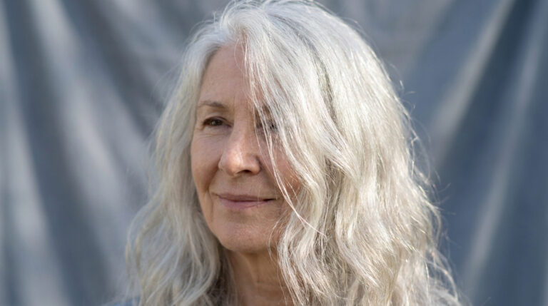 Portrait d'une femme senior aux longs cheveux gris ondulés sur fond gris.